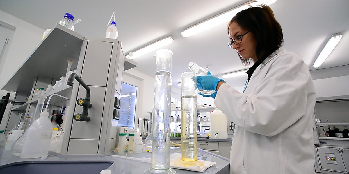 A woman in a lab coat pouring a solution from a flask into a graduated cylinder